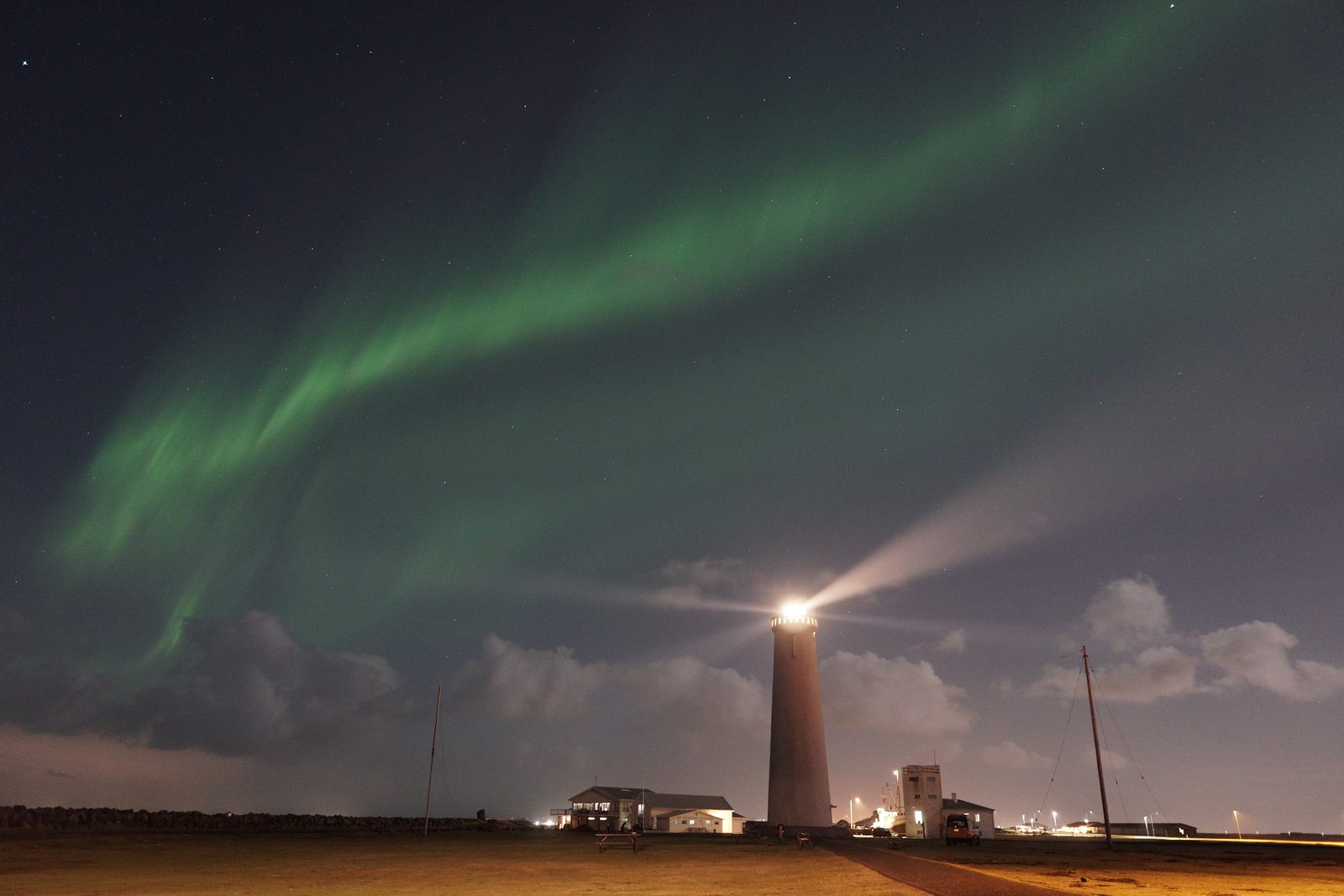 Green aurora arcing across a starry sky over a lighthouse whose beam cuts through low clouds