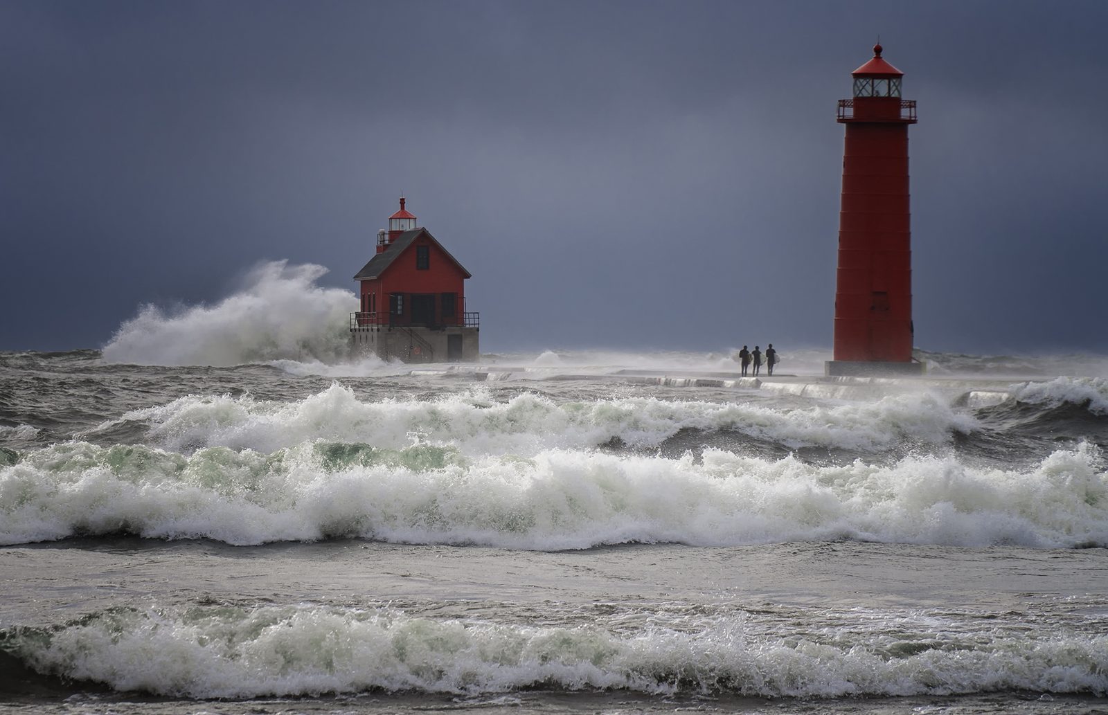 Storm waves breaking over a pier with two red lighthouses while three surfers carry their boards out